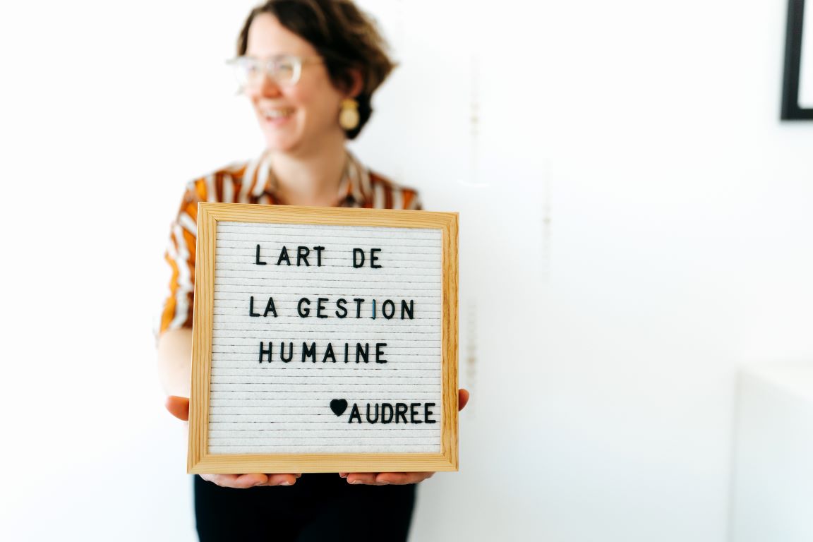 Une femme souriante, portant des lunettes et une chemise rayée, tient un tableau en bois où il est écrit 'L'art de la gestion humaine, Audree' en lettres noires sur fond blanc.